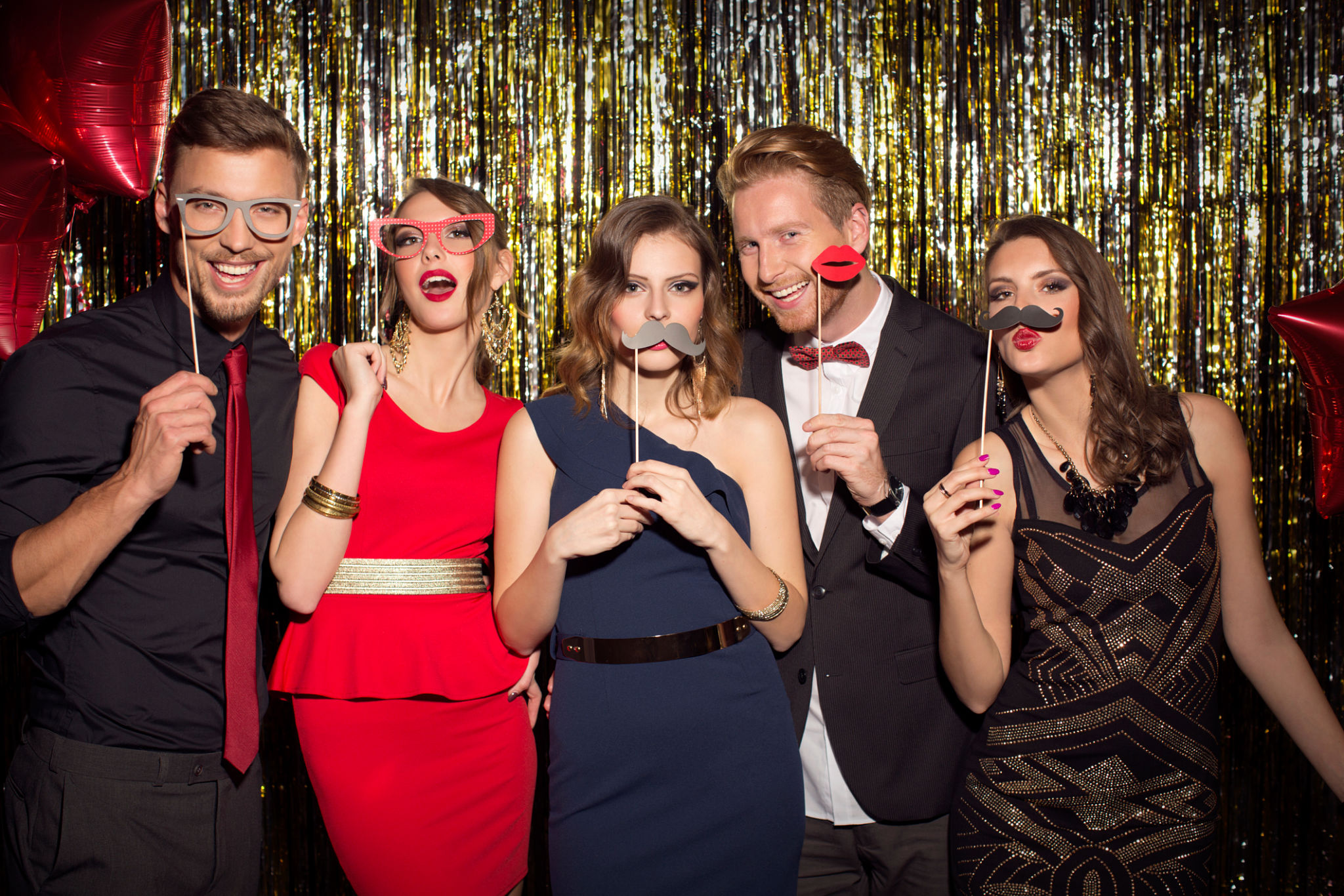 Young people wearing elegant clothes celebrating or having party in front of fringe curtain. They are holding photo booth fake props as moustaches, glasses and kiss lips and posing at camera.